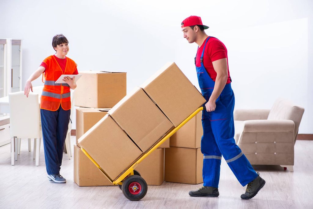 Professional movers in uniform handling cardboard boxes with a dolly during a residential move. One mover is guiding the process with a clipboard, ensuring an organized and efficient relocation.