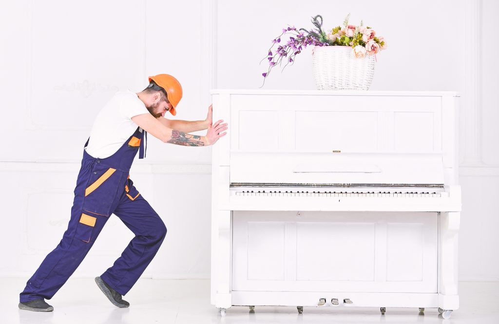 Professional piano mover in safety gear carefully pushing a white upright piano on wheels, with a floral arrangement on top, in a bright indoor setting.