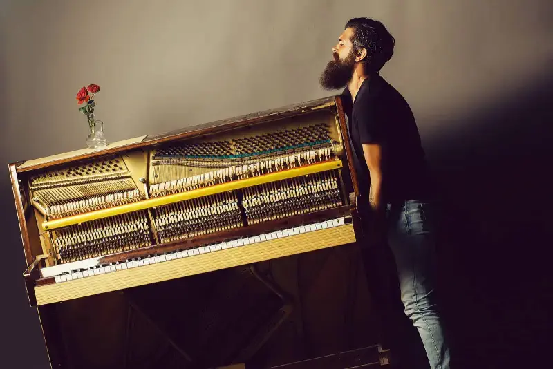 A bearded man in a black shirt and jeans carefully tilts an upright piano with its internal components exposed, highlighting the complexity of moving such an instrument. S.B Movers specializes in professional piano moving services, ensuring safe handling and transport of delicate pianos.