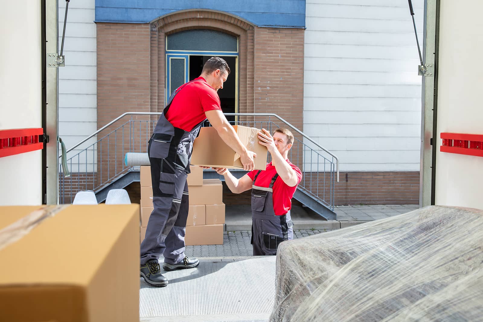 SB Movers team unloading moving boxes from a truck during a residential move. Two professional movers in red shirts and overalls carefully handling packed items outside a modern building.